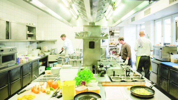 A commercial kitchen with stainless steel appliances, chefs preparing food, and various cooking utensils and ingredients on the counter. - Zum Vater Rhein (Monheim)