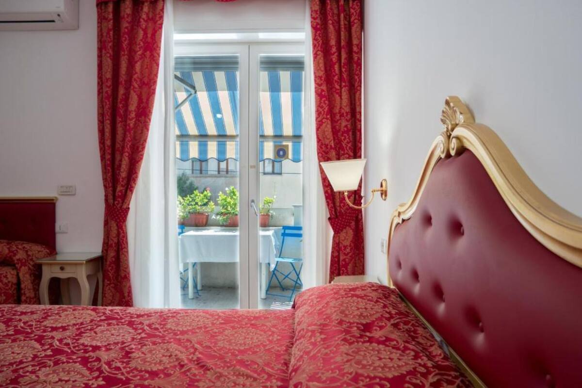 Ein Hotelzimmer mit rotem, gemustertem Bettbezug, einem roten, gepolsterten Kopfteil und Blick auf einen Außenbereich mit Tisch und Stühlen.