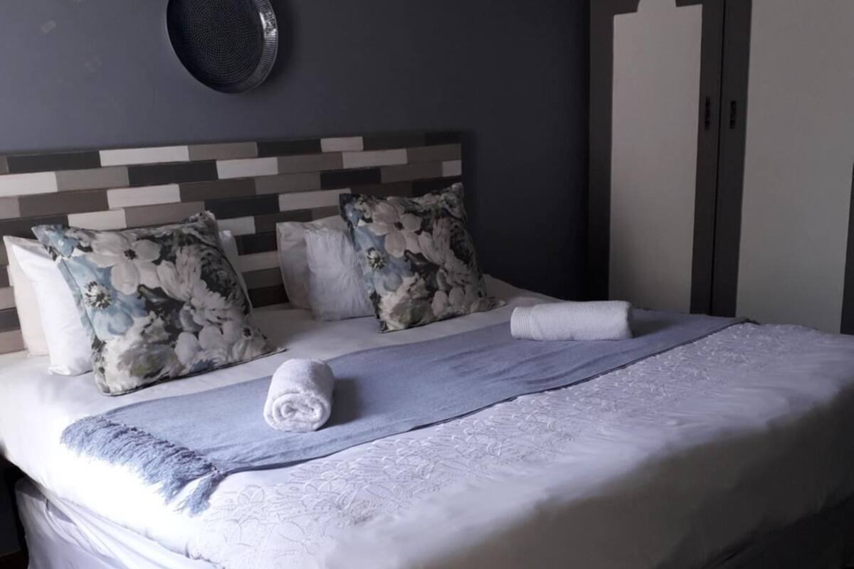 A neatly made bed with floral pillows and a white blanket, a round mirror on the wall, and a wardrobe with a geometric design.