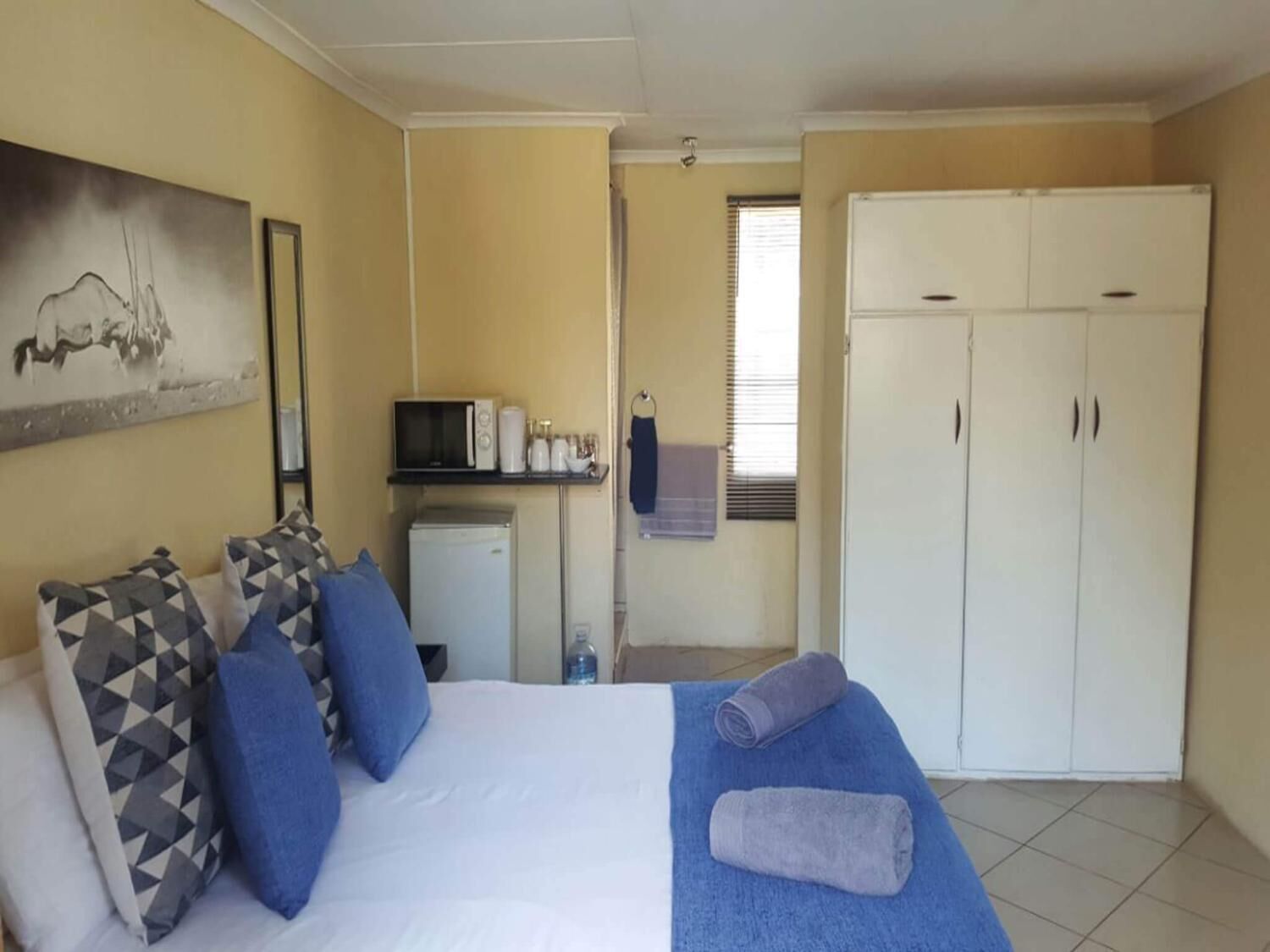 A bedroom with a bed, pillows, a blue blanket, a microwave, a small fridge, a wardrobe, and a painting on the wall.