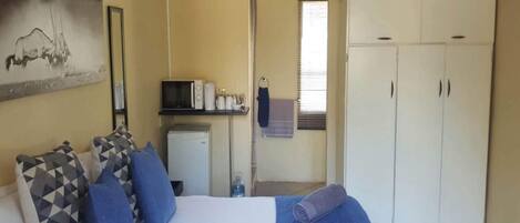 A bedroom with a bed, pillows, a blue blanket, a microwave, a small fridge, a wardrobe, and a painting on the wall.