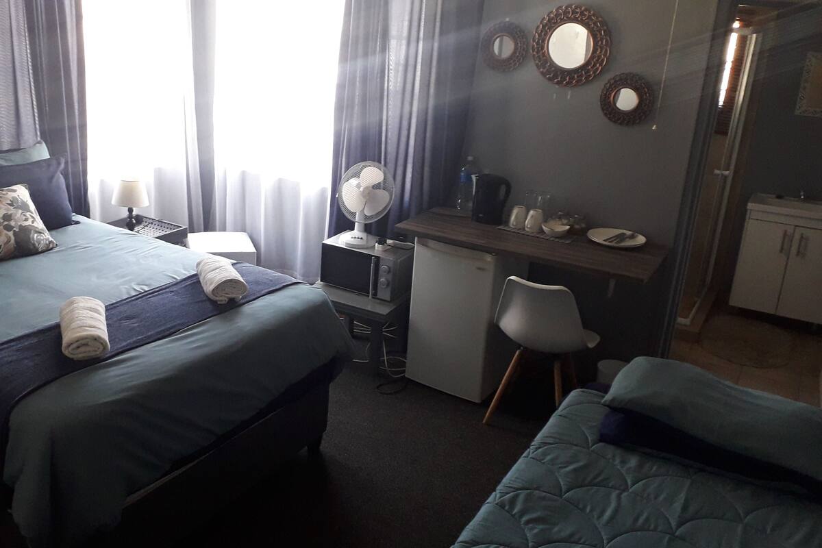A hotel room with a bed, a desk, a chair, a small kitchenette, and a mirror on the wall.