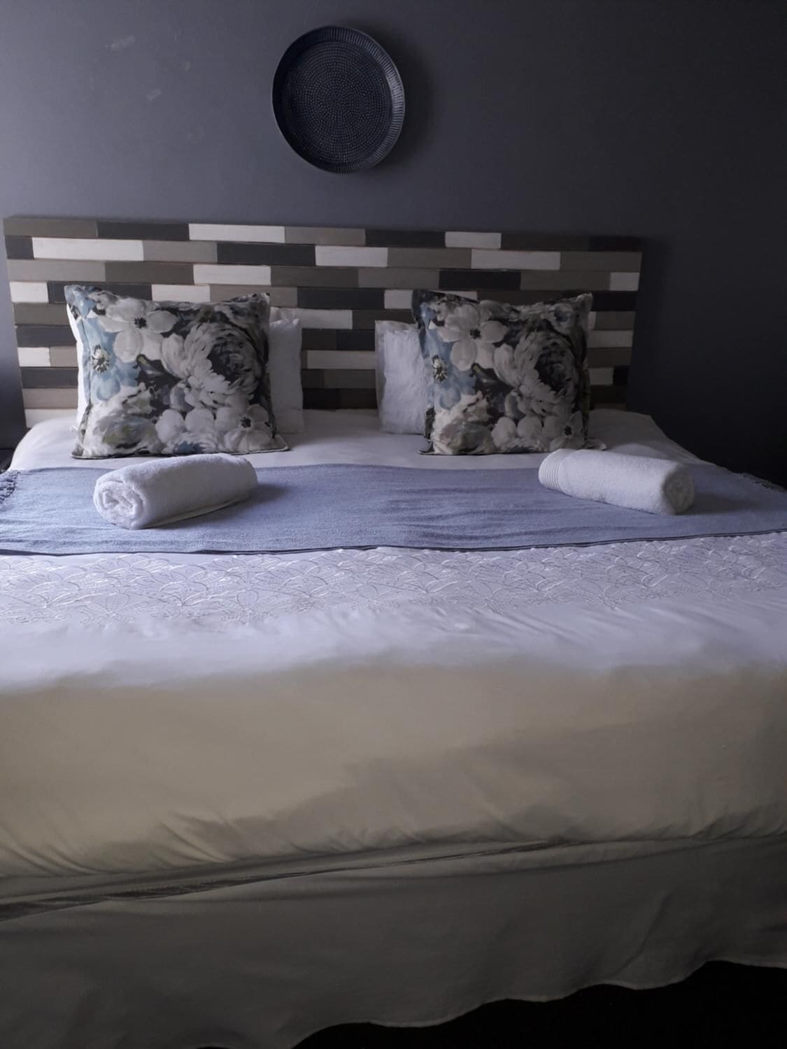 A neatly made bed with floral pillows and a rolled towel, a wall-mounted plate, and a striped headboard.