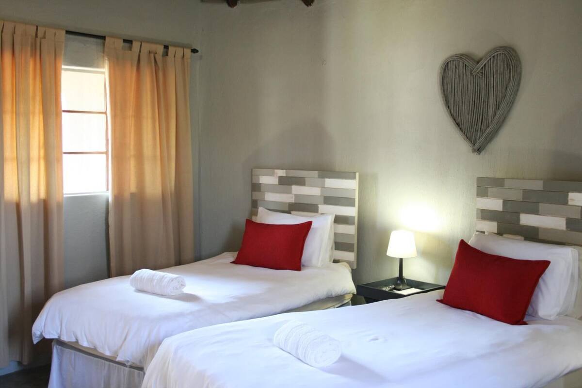 A hotel room with two beds, each with white bedding and red pillows, a heart-shaped wall decoration, and a window with curtains.