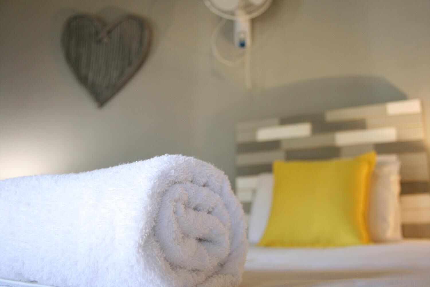 A neatly rolled white towel on a bed with a white and gray striped headboard, a yellow pillow, and a heart-shaped decoration on the wall.