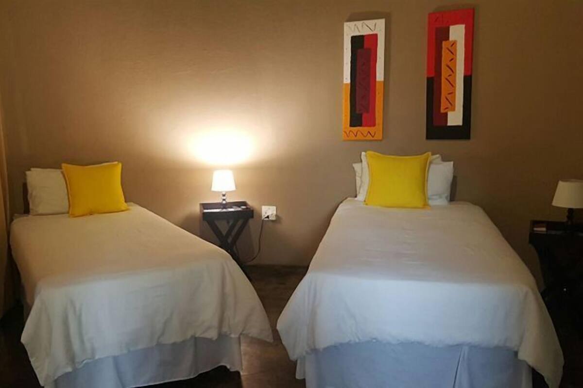 A hotel room with two beds, each with white bedding and a yellow pillow. There is a nightstand with a lamp between the beds. The walls are beige, and there are two pieces of artwork above the nightstand.