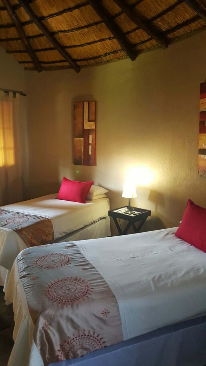 A room with two beds, each with a white bedspread and a red pillow. There is a small table with a lamp between the beds.
