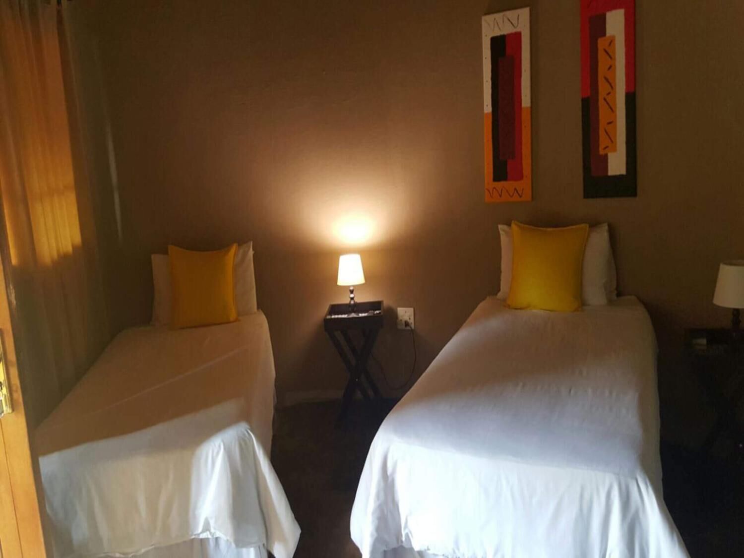 A hotel room with two single beds, each with a white bedspread and a yellow pillow. There is a small table with a lamp between the beds, and a wall-mounted artwork above the table.