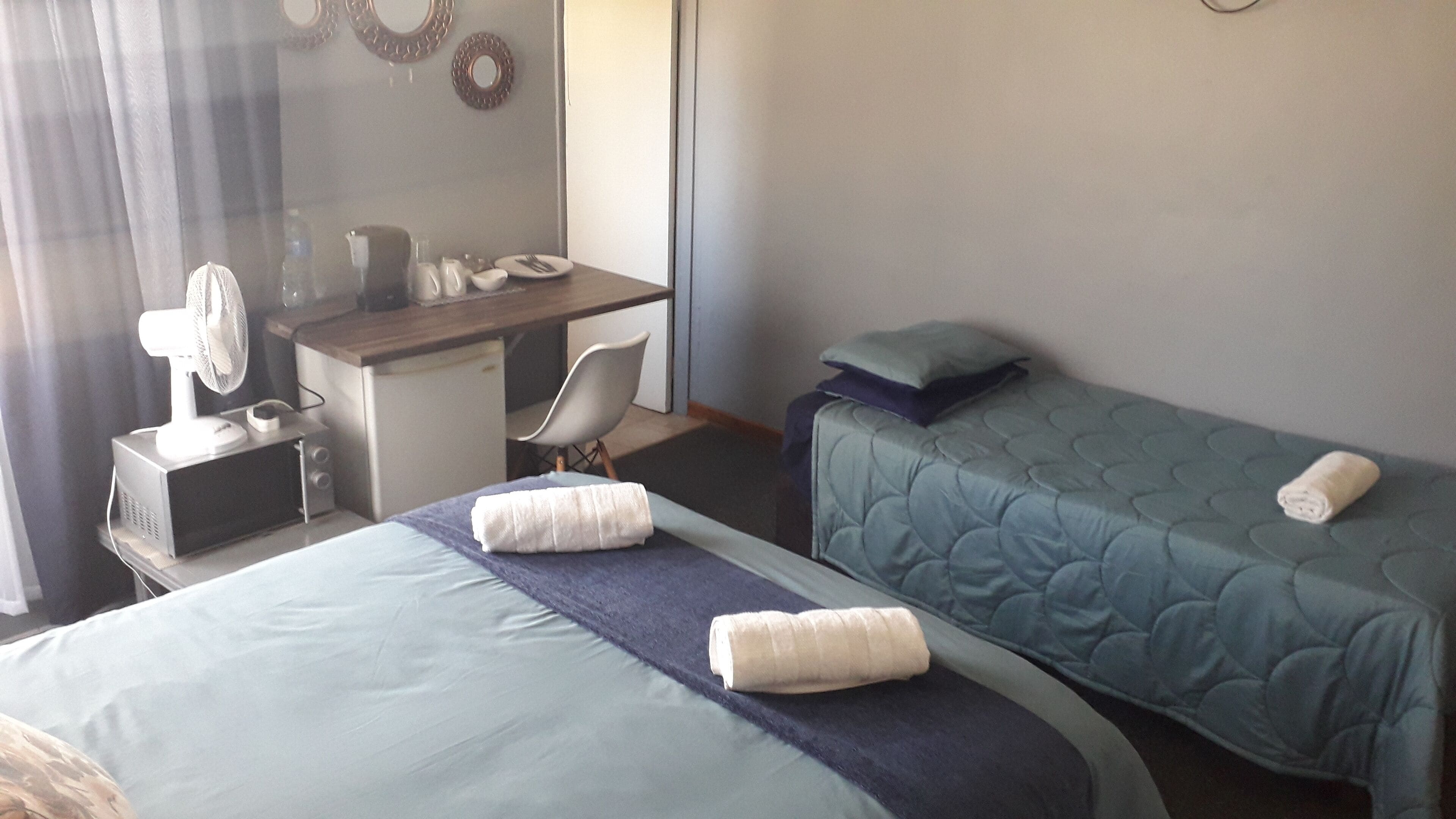 Habitación de hotel con dos camas, un escritorio, una silla, una mesita con un ventilador y una ventana con cortinas.
