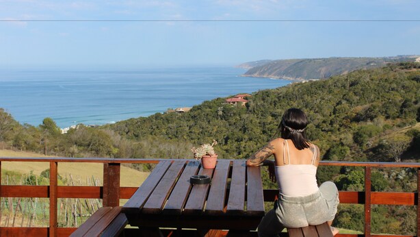 Mulher sentada à mesa de madeira com um vaso de planta, apreciando a paisagem costeira com colinas e vista para o mar.