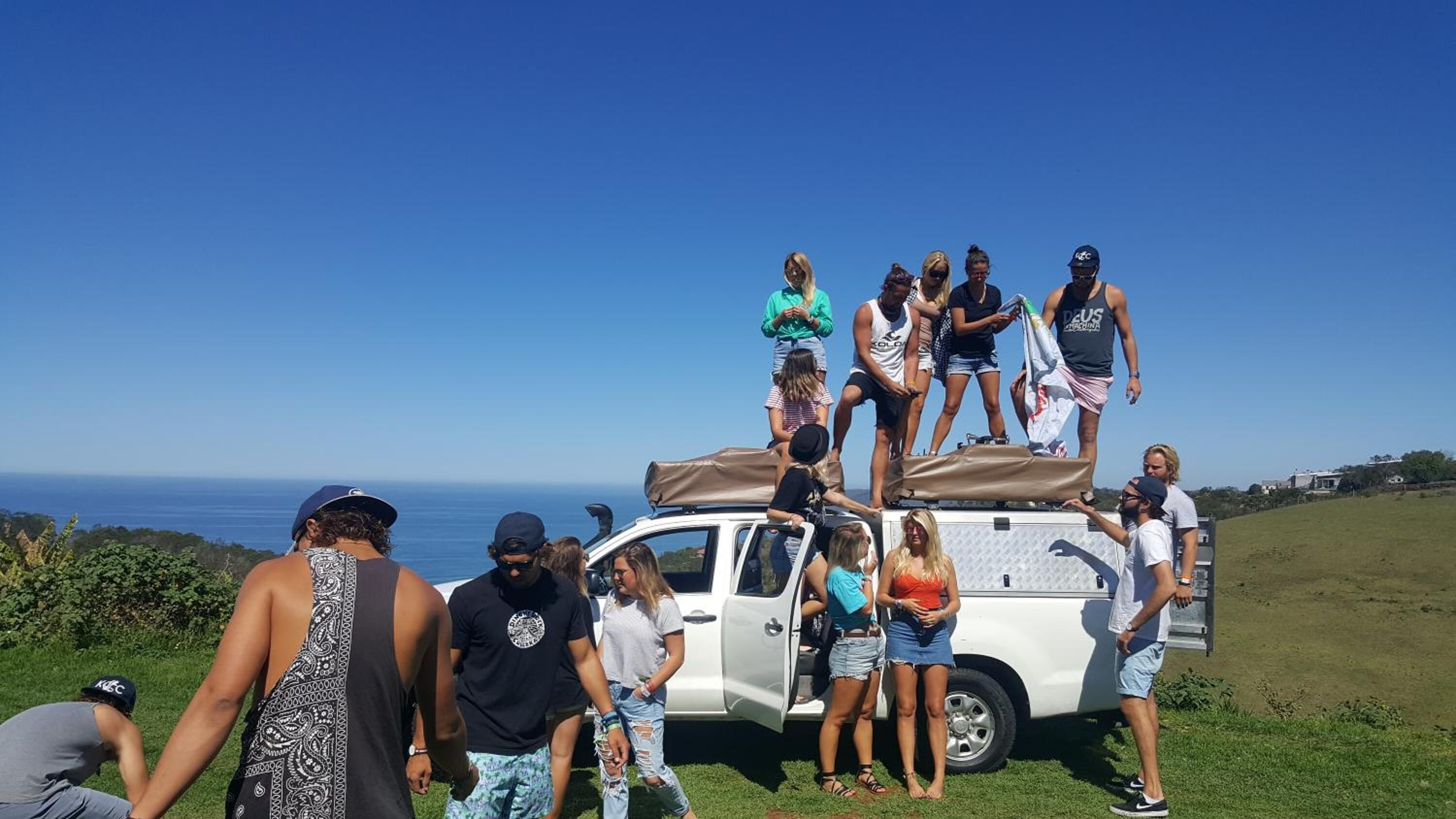 A group of people standing around a white vehicle on a grassy area with a clear blue sky and ocean in the background.
