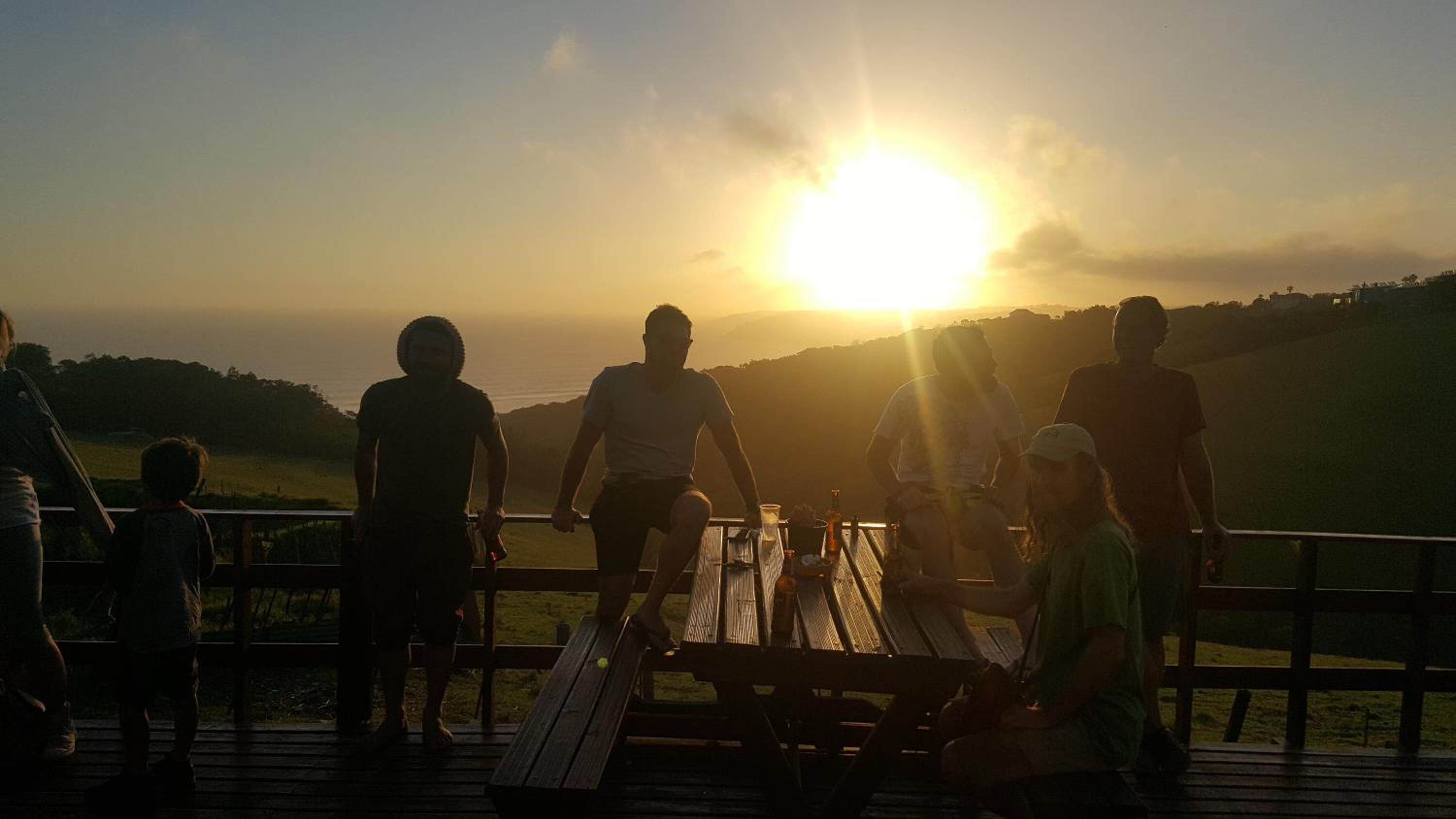 A group of people on a wooden deck overlooking a scenic landscape with a sunset in the background.