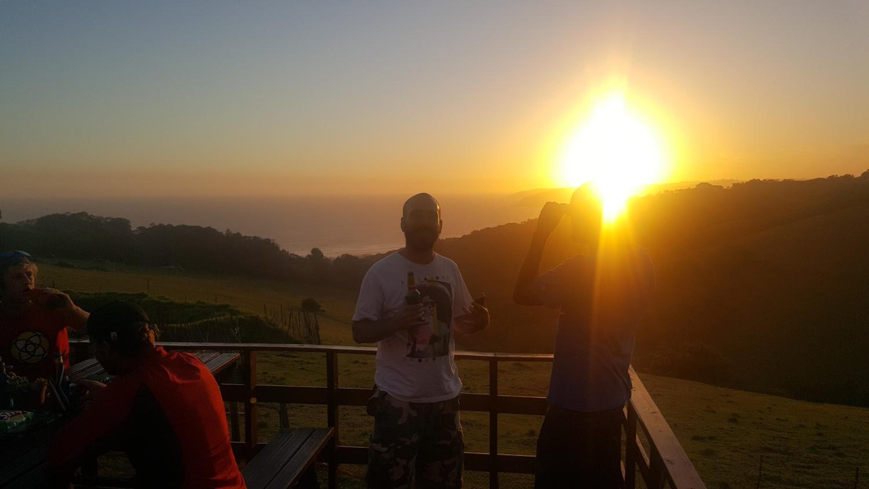 Vista del atardecer desde una terraza de madera con baranda, sobre un paisaje cubierto de hierba.