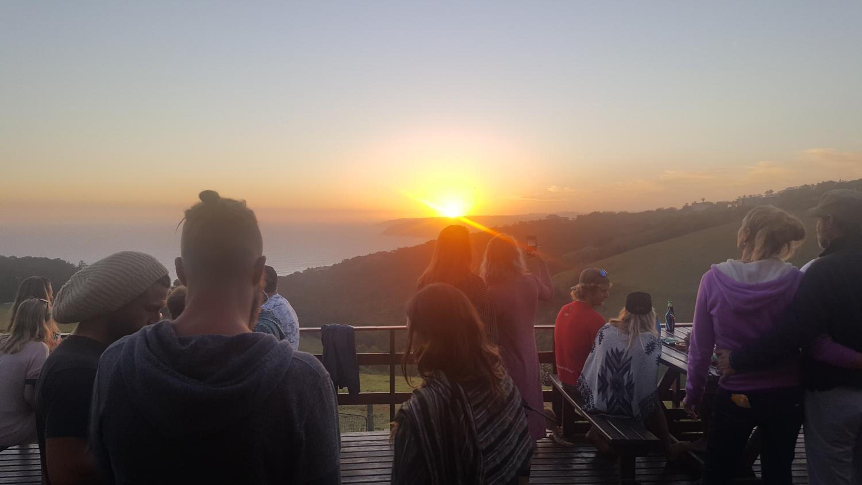 Un grupo de personas en una terraza de madera con vistas a la puesta de sol y al paisaje.