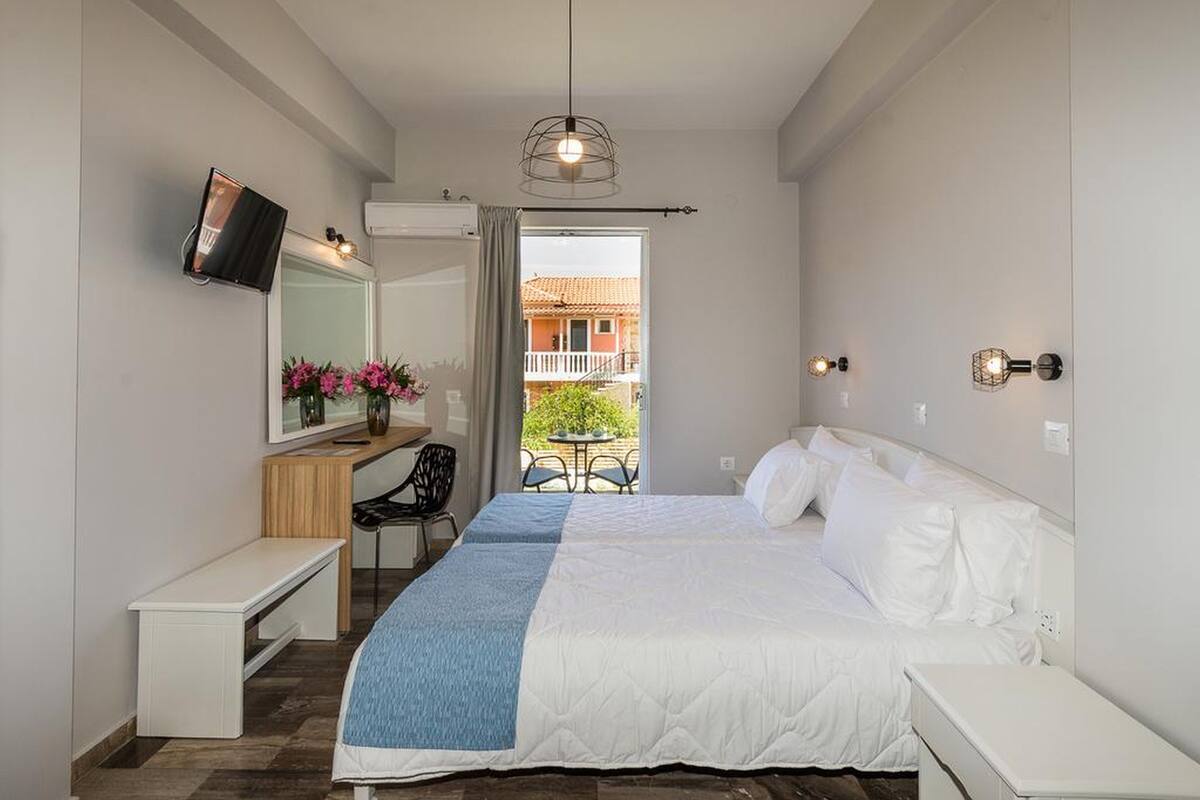 A bedroom with a bed, a desk, a chair, a TV, and a balcony with a view of buildings.
