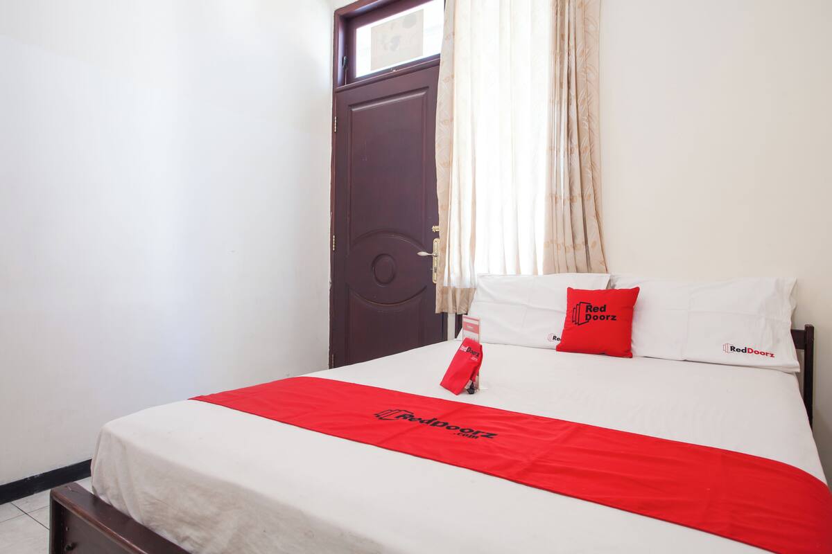 A hotel room with a bed, a red and white bedspread, a red pillow, and a door with a window.