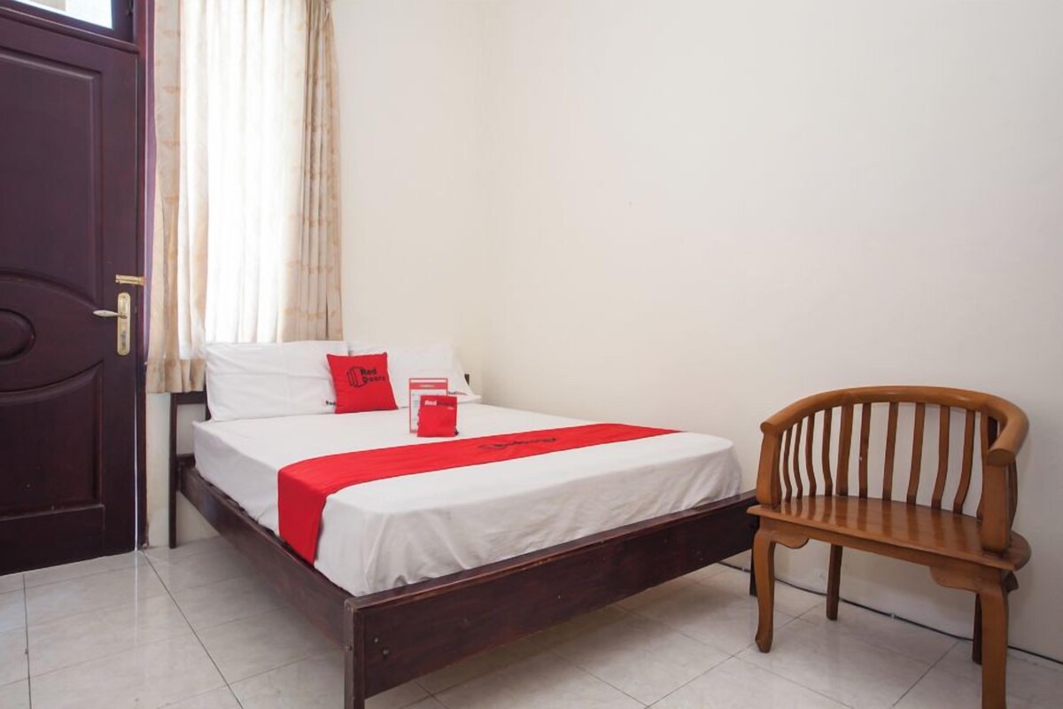 A single bed with a wooden frame, white bedding, and a red throw. A wooden chair is beside the bed.