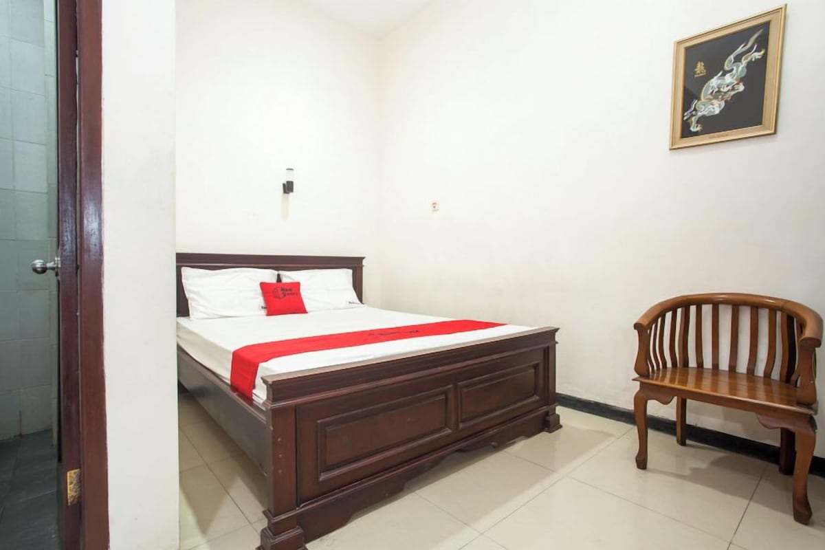 A bedroom with a wooden bed, a red bedspread, a wooden bench, and a framed artwork on the wall.