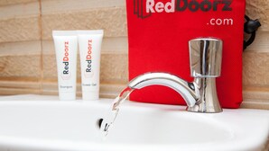 A bathroom sink with a chrome faucet and running water, two RedDoorz shampoo and shower gel tubes, and a red RedDoorz bag hanging on the wall.