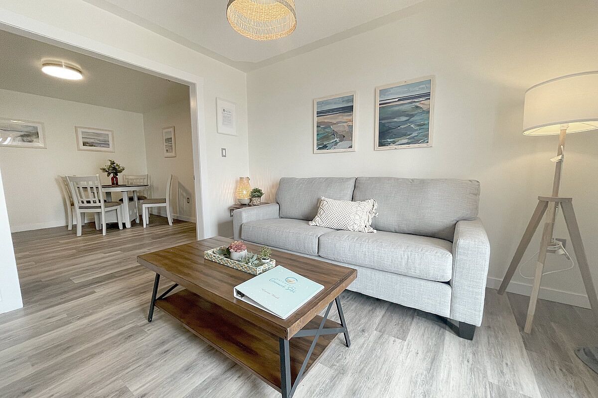 A modern living room with a grey sofa, a wooden coffee table, and a dining area with white chairs.
