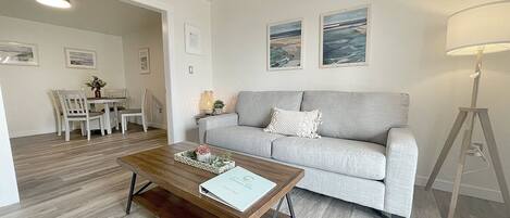 A modern living room with a grey sofa, a wooden coffee table, and a dining area with white chairs.
