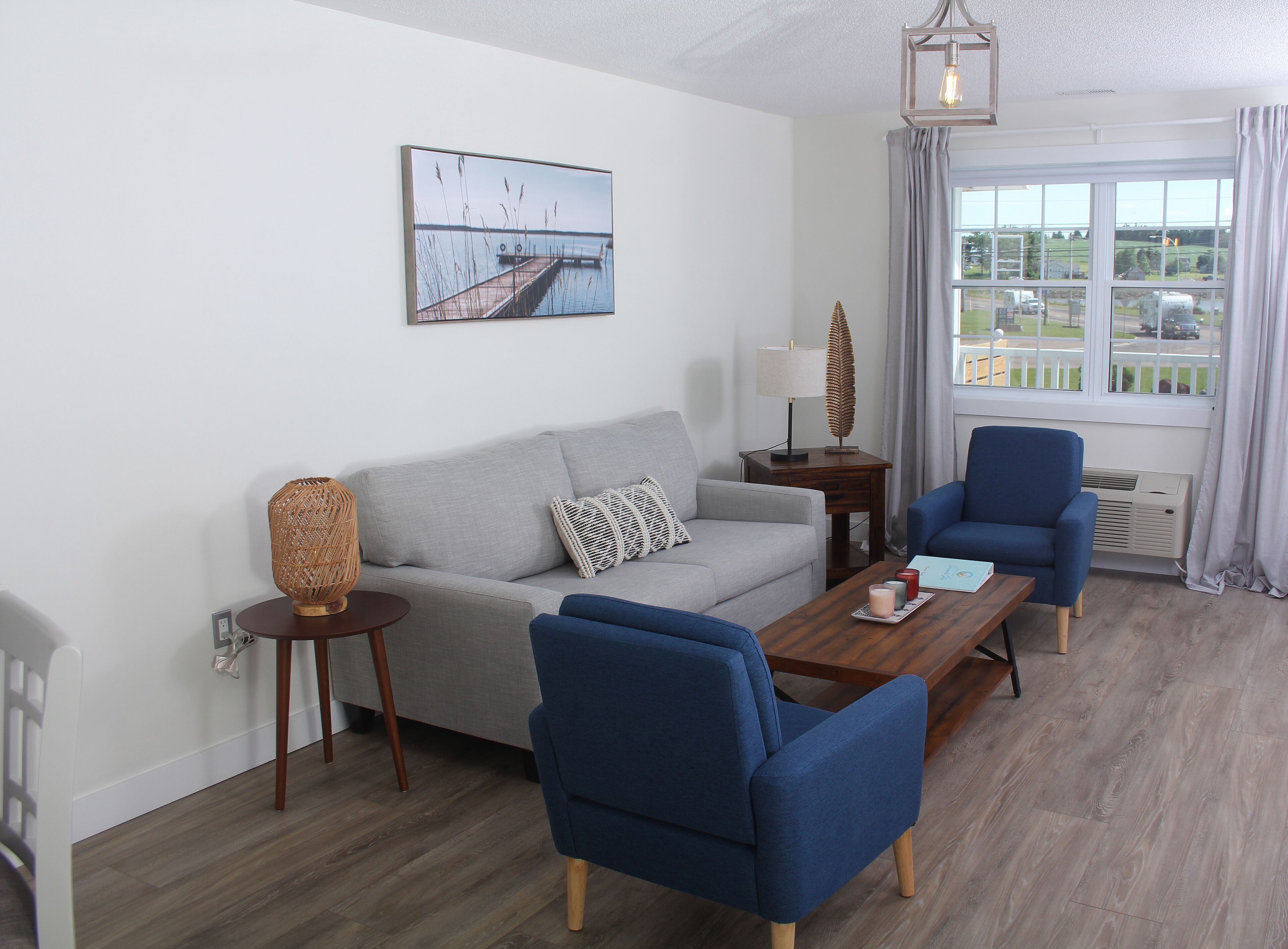 Una sala de estar moderna con un sofá gris, sillones azules, una mesa de centro de madera y una lámpara decorativa. En la pared, un cuadro de un muelle y agua.