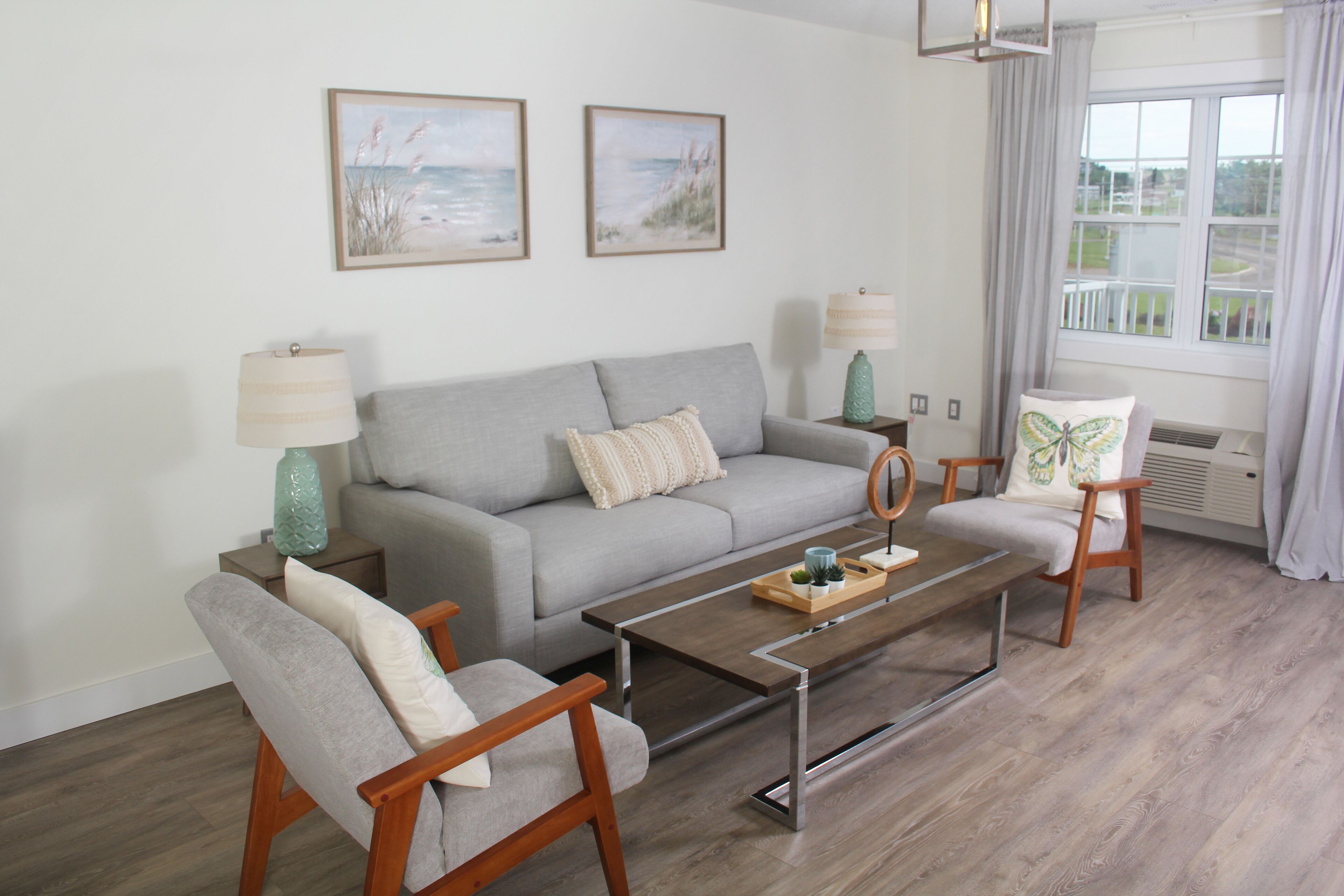 A modern living room with a grey sofa, two armchairs, a coffee table, and a side table with lamps. There are two framed pictures on the wall, a window with curtains, and a view of the outdoors.
