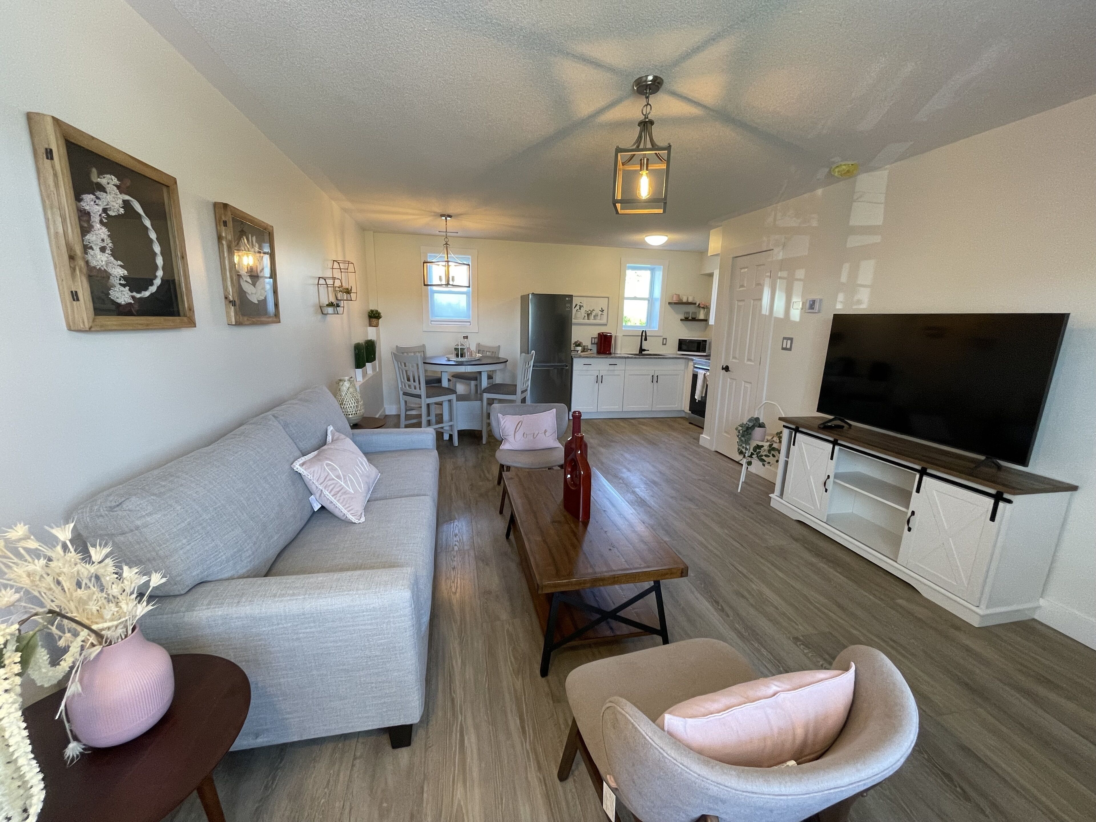 A modern living room with a grey sofa, a wooden coffee table, and a TV on a wooden stand. There is a kitchen in the background with white cabinets and a dining area.