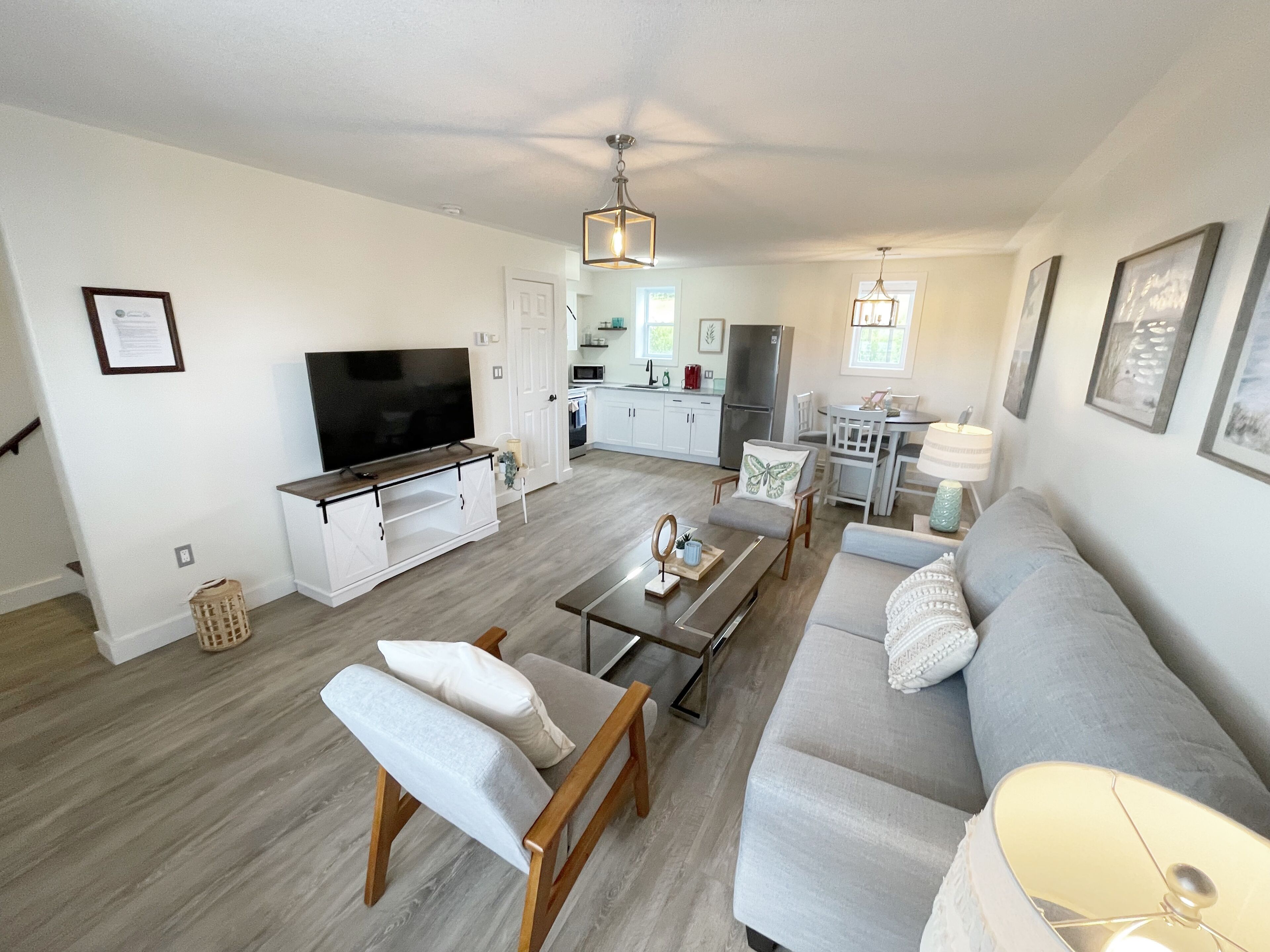 A modern living room with a sofa, armchairs, a coffee table, and a TV. There is a kitchen in the background with a refrigerator, microwave, and dining area.