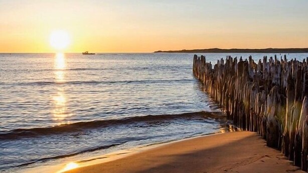 Une plage avec une jetée en bois qui s’avance dans l’eau, un bateau au loin et un coucher de soleil à l’horizon.
