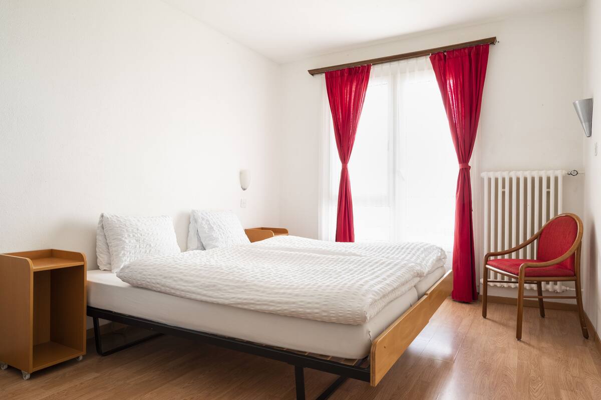 Una cama individual con estructura de madera, una mesita de noche de madera, una silla roja y un radiador.