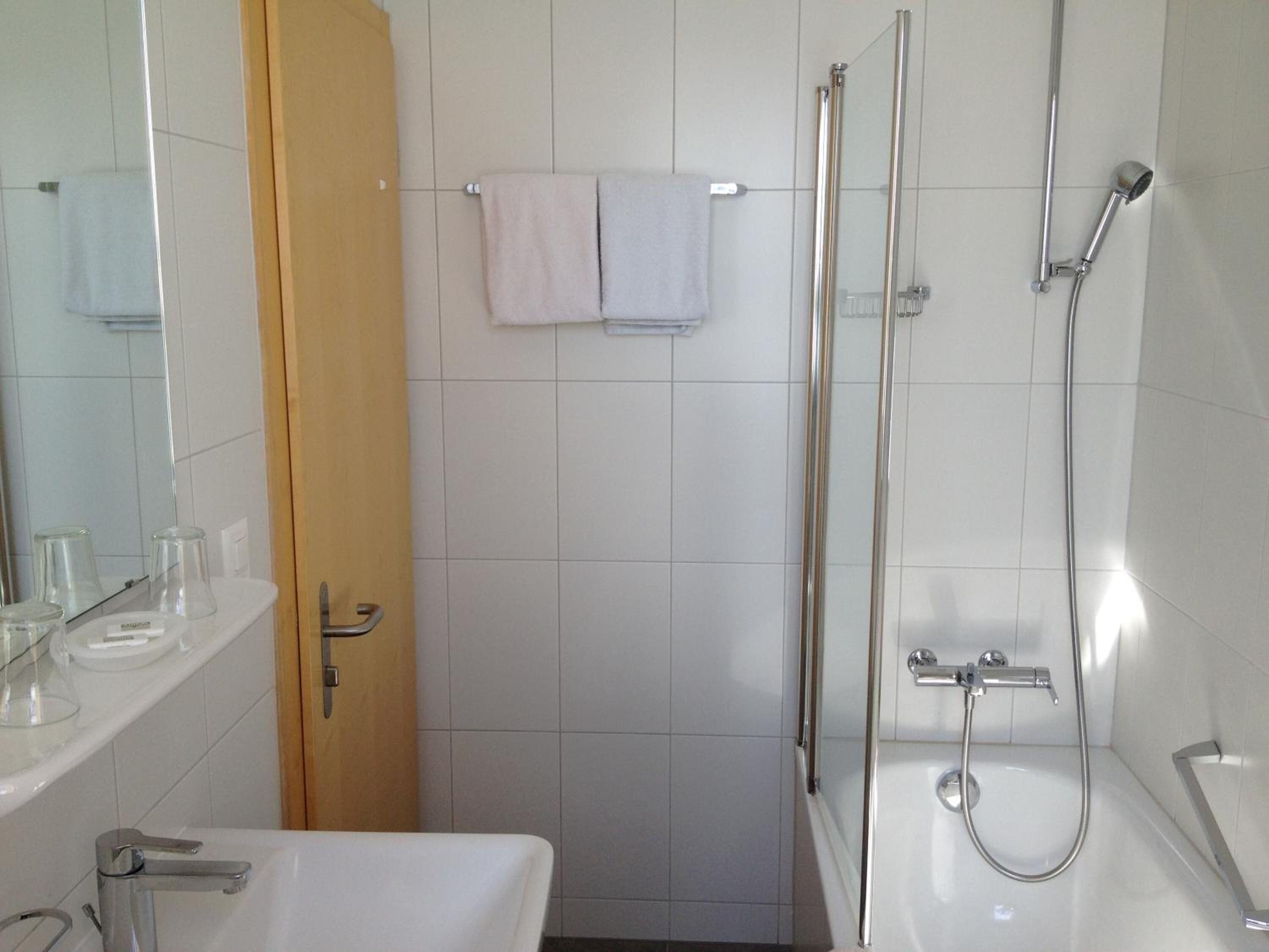 Een badkamer met een witte wastafel, een spiegel, een handdoekhouder met twee handdoeken, een douche met een handdouche en een douchecabine van glas.