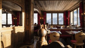A wooden interior with tables and chairs, large windows with curtains, and a view of snow-capped mountains. - Hotel Schwarzsee (Zermatt)