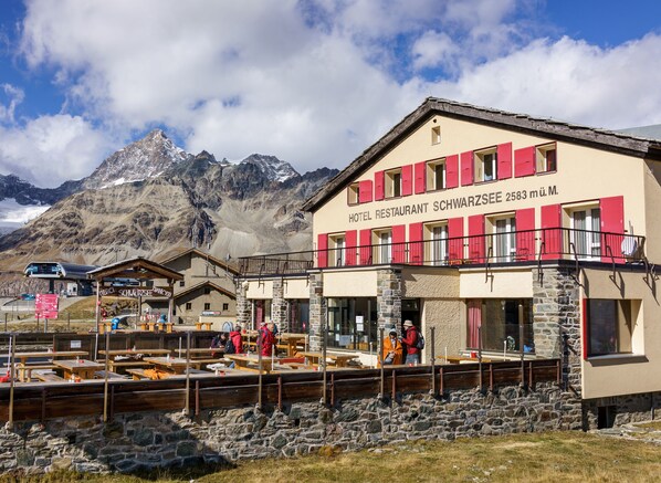 Un hotel con montañas al fondo, con fachada de piedra, contraventanas rojas y mobiliario exterior.