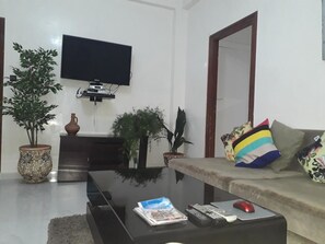 A living room with a flat-screen TV mounted on the wall, a sofa with colorful cushions, a coffee table with remote controls and a magazine, and potted plants. - Train station apartment Casa Voyageur (Casablanca)