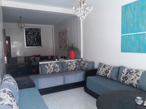 A modern living room with a sectional sofa, a coffee table, and a chandelier. There is a potted plant on a shelf and artwork on the walls. - Train station apartment Casa Voyageur (Casablanca)
