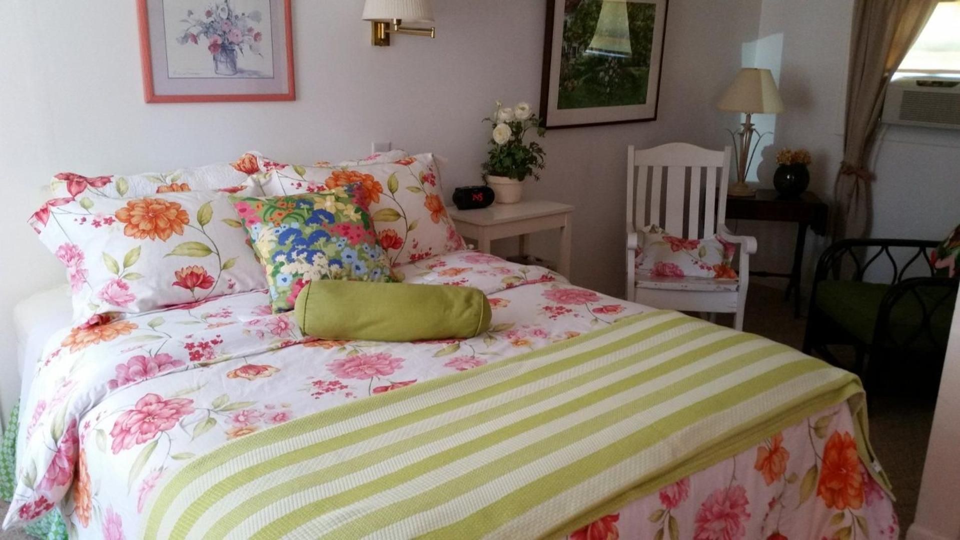 A bed with floral bedding, a green pillow, and a striped blanket. There is a chair with a floral cushion, a small table with a lamp, and a framed picture on the wall.