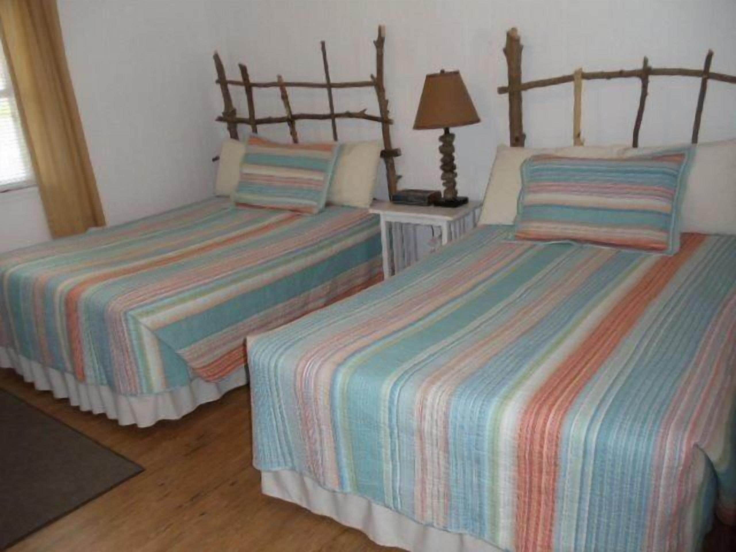Two single beds with striped bedding, a wooden headboard, a bedside table with a lamp, and a window with curtains.