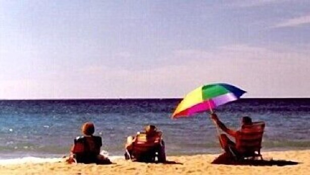 Three people sitting on the beach with a colorful umbrella. - Bay Port Lodging (Frankfort)