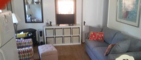 A living room with a grey sofa, a television on a stand, a white cabinet, and a patterned ottoman.