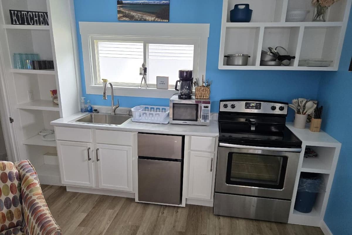 A compact kitchen with white cabinets, a stainless steel refrigerator, a microwave, an oven, and a window above the sink.