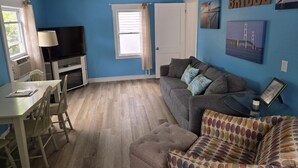 A living room with a blue wall, a grey sofa, a white table with chairs, a television, and a window with curtains. - Bay Port Lodging (Frankfort)