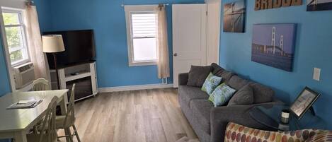 Una sala de estar con pared azul, sofá gris, mesa blanca con sillas, televisor y ventana con cortinas.