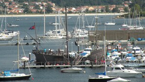 A marina with various boats docked, a pier with people, and a backdrop of houses and hills. - Hotel Rompeolas (Baiona)