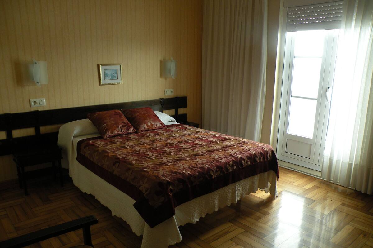 A bedroom with a wooden floor, a bed with a floral bedspread, a nightstand, a bench, a wall-mounted light, a framed picture, and a window with curtains.