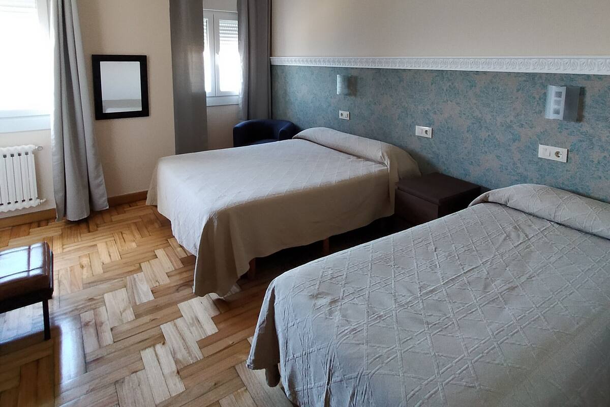 A hotel room with two beds, a wooden herringbone floor, a radiator, and a window with curtains.