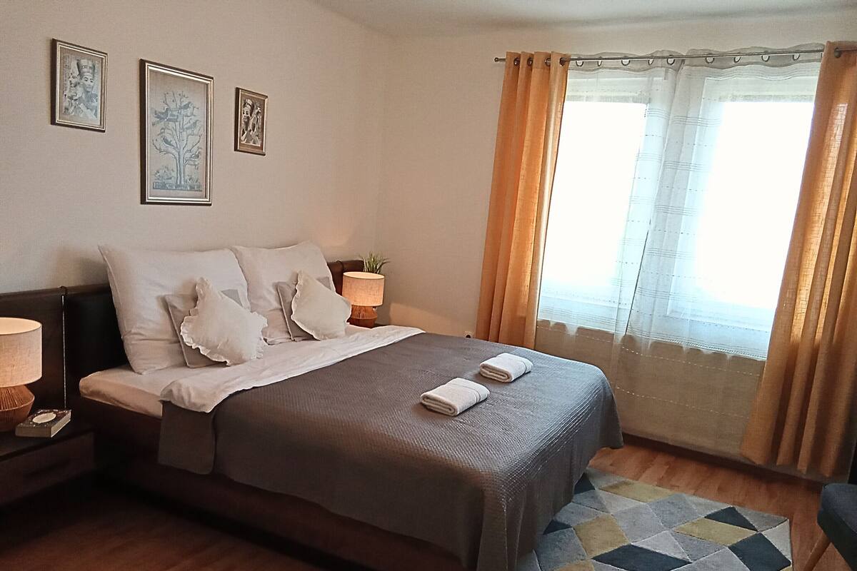 A neatly made bed with pillows and a blanket, a bedside lamp, and a window with curtains.