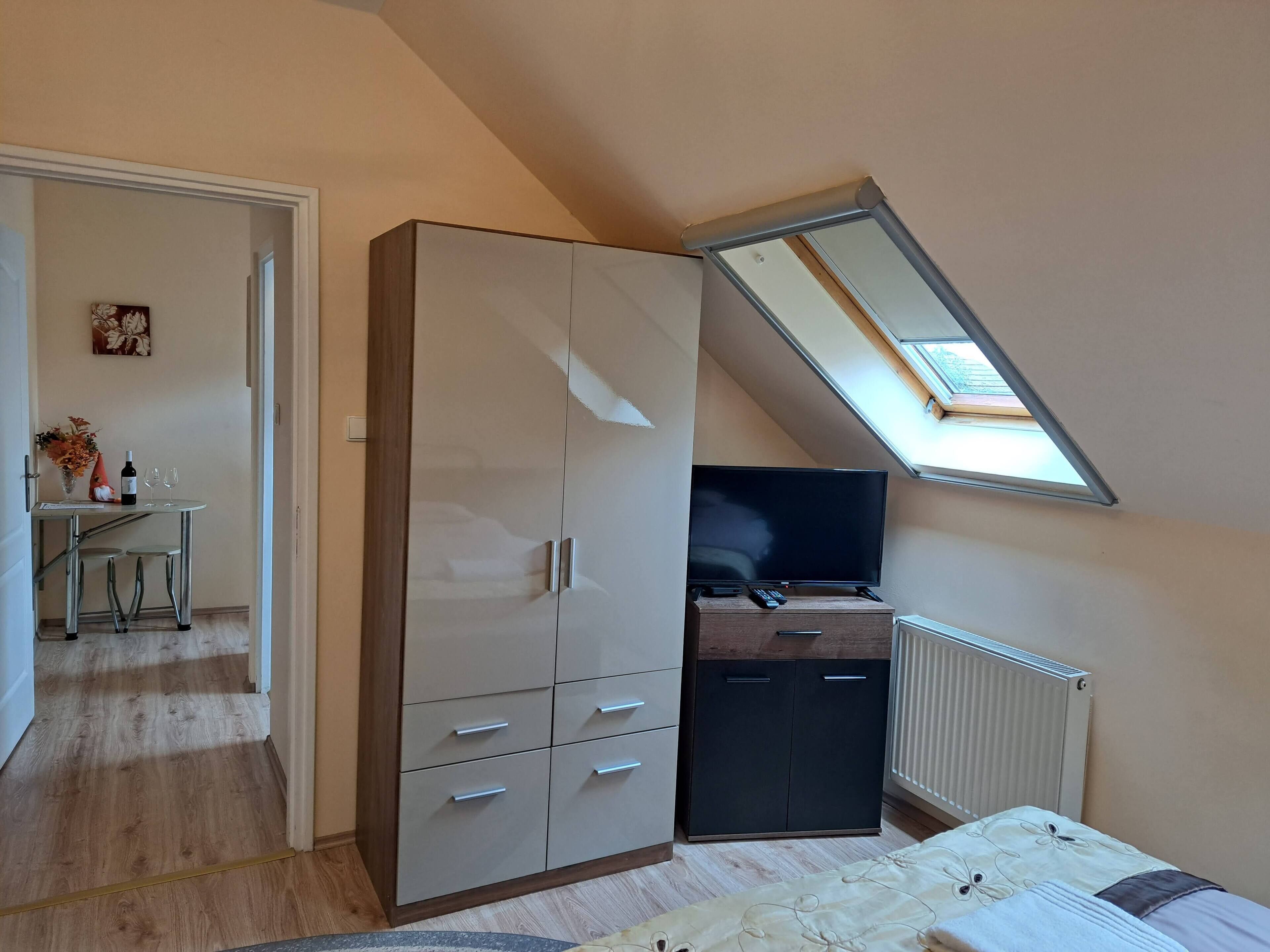A bedroom with a sloped ceiling, a bed, a wardrobe, a desk with a chair, and a radiator.