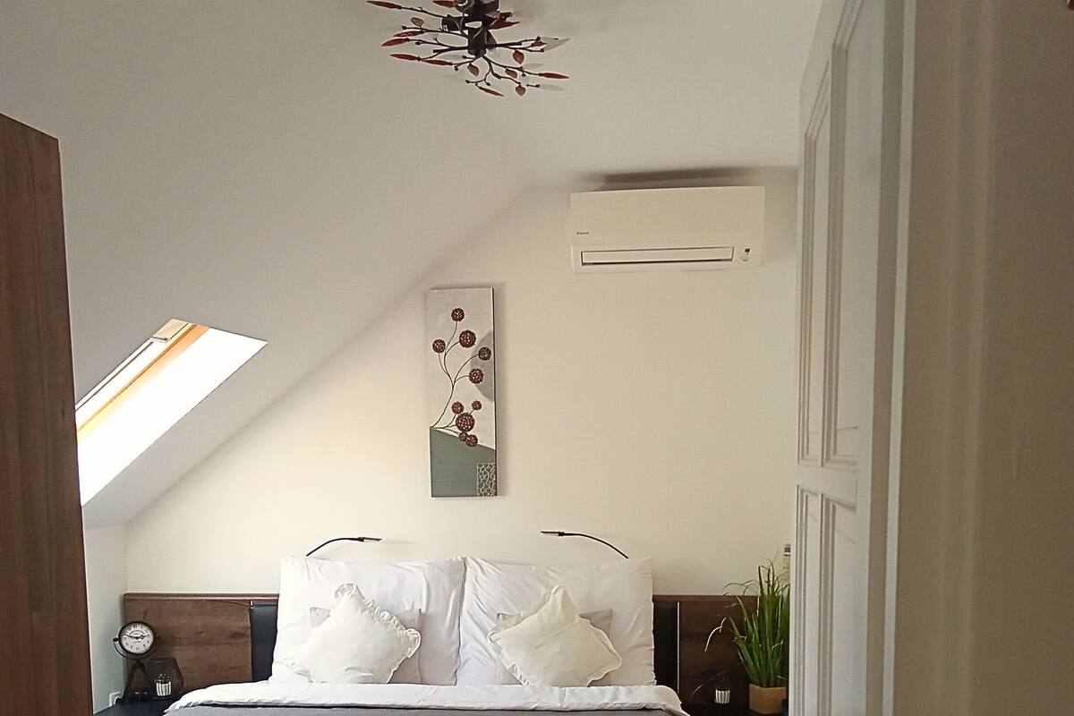 A bedroom with a bed, a nightstand, a wall clock, a ceiling fan, a skylight, and a painting.