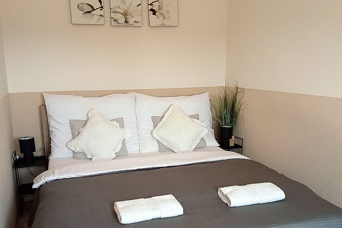 A neatly made bed with white pillows and a grey blanket, a small bedside table with a lamp, and three framed floral artworks on the wall.